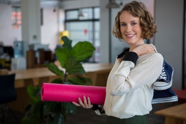 Smiling Female Designer Holding Yoga Mat and Sneakers in Office - Download Free Stock Photos Pikwizard.com