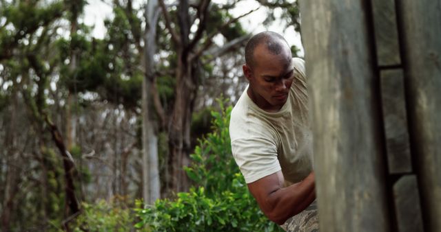 This photo depicts a carpenter with a shaved head working on an outdoor wooden structure surrounded by trees and greenery. Due to the detailed environment, it suggests the carpenter is engaged in constructing something like a hut or cabin at some camp or park area. Perfect for promoting outdoor work, construction projects, or natural building techniques.