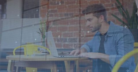 Focused young man typing on laptop in brick-walled cafe with coffee, greenery and daylight