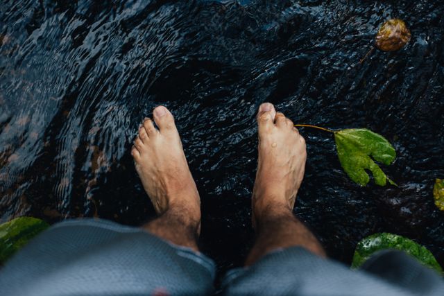 Image depicts a man standing with bare feet in a calm stream, highlighting natural relaxation and tranquility. Useful for concepts related to stress relief, outdoor activities, connection with nature, and environmental awareness.
