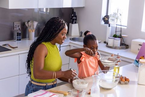 African american mother and daughter baking together, sifting flour in modern kitchen