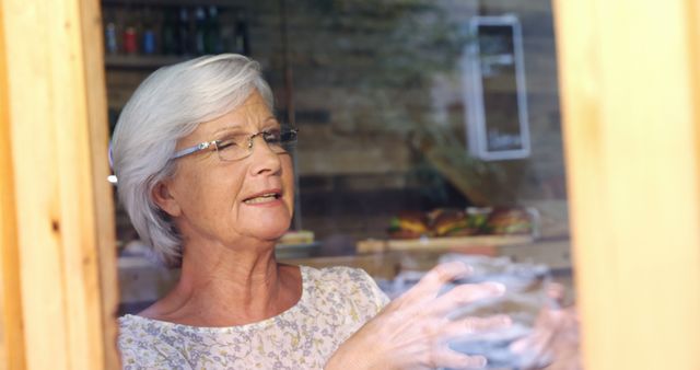 A senior Caucasian woman looks thoughtfully out of a window, with copy space. Her expression suggests she may be reminiscing or enjoying a moment of quiet reflection.