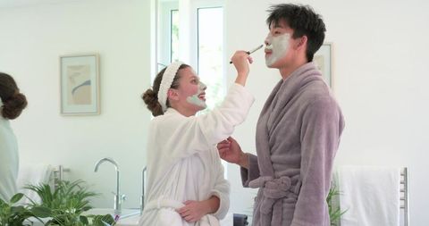 Couple enjoying at-home skincare routine applying facial masks in bright bathroom