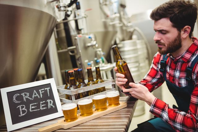 Young brewer examining craft beer bottles in brewery. Ideal for use in articles or advertisements related to brewing industry, craft beer production, quality control in breweries, and beer tasting events. Suitable for illustrating topics on artisanal beer, small-scale brewing businesses, and beer-making processes.