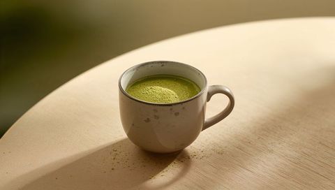 Speckled ceramic mug holding bright matcha powder on round wood table in soft light
