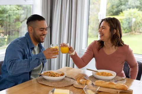 Couple toasting with orange juice over cozy pasta dinner at bright dining table