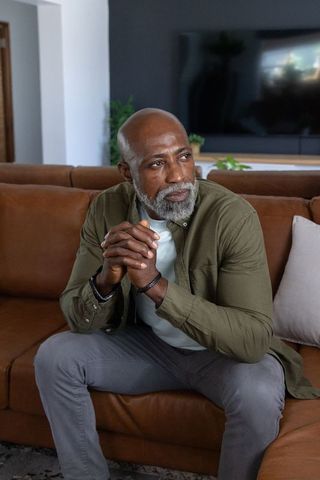 Contemplative senior african american man sitting on brown sofa gazing at tv in modern cozy living r