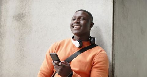 Young African American Man Leaning on Concrete Wall, Smiling While Holding Smartphone