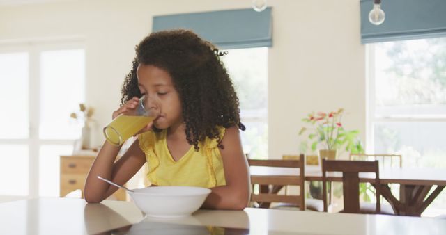 Young girl in yellow dress drinking juice while sitting at kitchen table for breakfast. Ideal for concepts of family routines, healthy eating, and morning activities at home. Suitable for use in advertisements, educational materials, or health and nutrition campaigns.