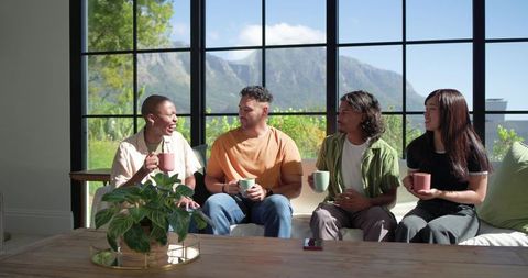 Diverse friends chatting on sofa with mugs and big window mountain view, casual living room