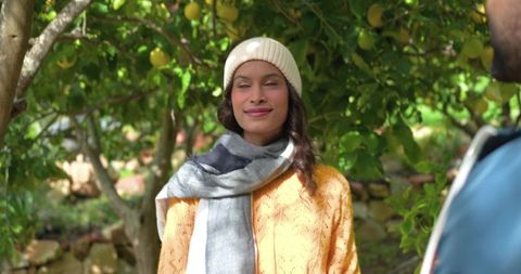Smiling woman under lemon tree with partner in sunlit orchard wearing beanie sweater