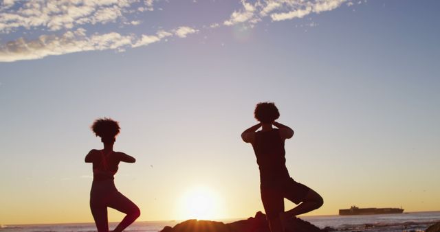 Silhouettes of two individuals performing yoga poses on a beach during sunrise, highlighting a serene and peaceful atmosphere. Ideal for concepts related to mindfulness, fitness, wellness activities outdoor, starting the day with meditation, and promoting relaxation and healthy lifestyle. Can be used for articles, blog posts, and social media promoting yoga retreats, beach workouts, or mindfulness practices.