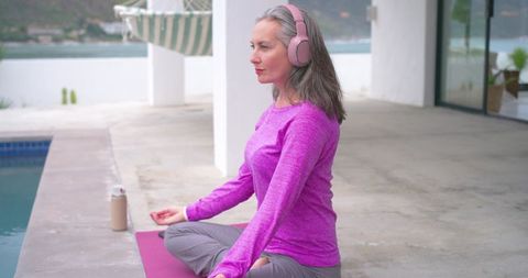 Mature woman practicing mindful meditation by pool wearing headphones and activewear