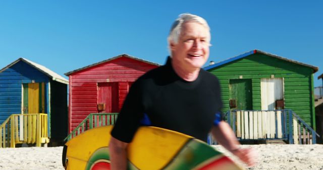 A senior Caucasian man is smiling as he carries a surfboard on a sunny beach with colorful beach huts in the background. His active lifestyle and joy are evident as he enjoys a day of surfing by the sea.