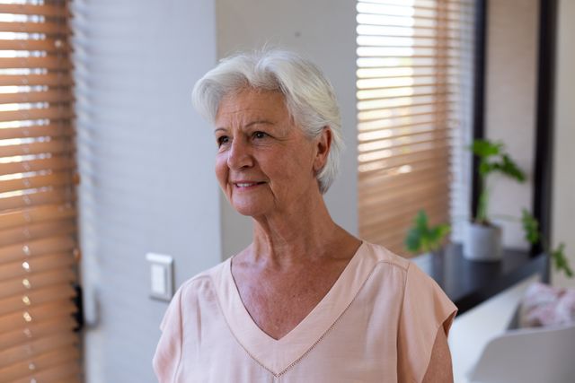 Senior caucasian woman standing in living room, gazing out window with a thoughtful expression. Natural light illuminates the room, highlighting indoor plants and a cozy home environment. Ideal for use in retirement lifestyle promotions, senior living advertisements, and articles on aging gracefully.