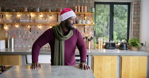African American man wearing Santa hat and scarf leaning on rustic kitchen island