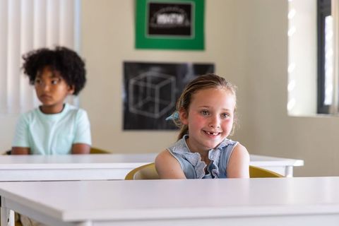 Smiling schoolgirl sitting at desk in bright classroom with diverse classmates