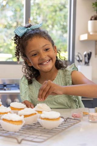Joyful girl decorating cupcakes with sprinkles in sunny home kitchen