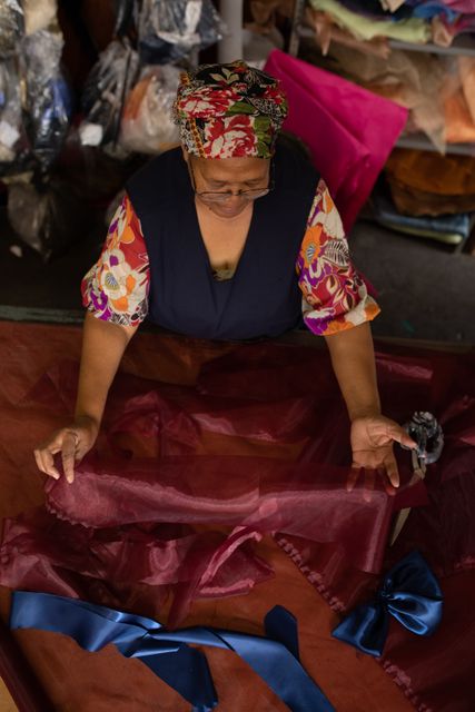 High angle front view of a biracial woman standing at a table working with purple fabric at a hat factory. In the background bales of material can be seen in storage.
