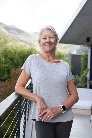 Senior woman standing on balcony wearing smartwatch and light gray top smiling on terrace