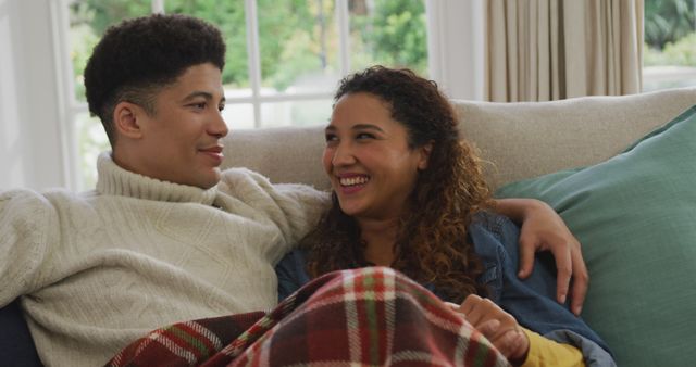 Young mixed race couple enjoying intimate moment while cuddling on a couch wrapped in a blanket. Perfect for depicting themes of love, companionship, interracial relationships, comfort, and domestic life.