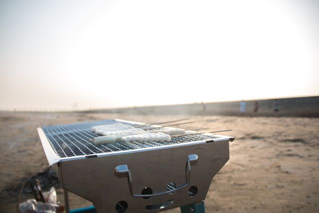 Close shot of food grilling on metal grill at beach on sunny day. Background shows sandy shore and distant horizon. Ideal for illustrating outdoor, beach activities, summer vacations, and food preparation in nature settings.