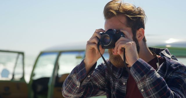 Young man in casual clothing using camera to take photos outdoors with blurred vehicle background. Ideal for use in lifestyle, travel, photography tutorials, and outdoor adventure promotions.