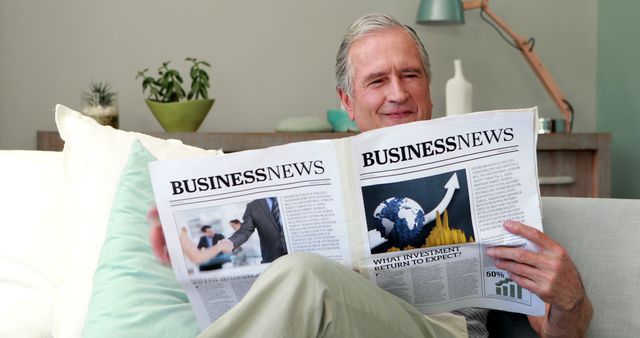 Senior man sitting comfortably at home, engaging in leisure activity of reading business newspaper. Suitable for use in financial advisories, lifestyle articles targeting retired individuals, or promotions of investment news platforms.
