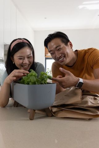 Young couple tending indoor herb planter on sunlit kitchen island, smiling and cooking