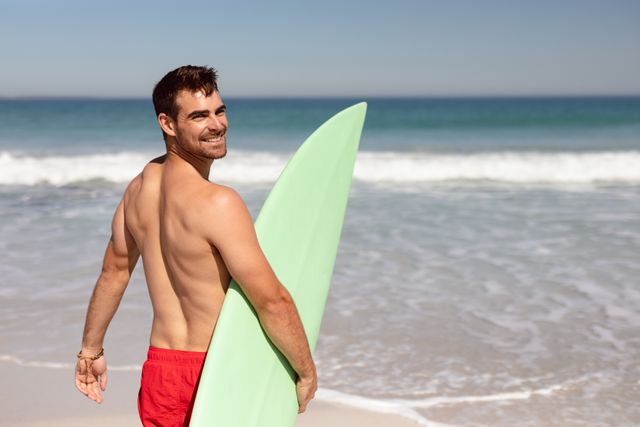 Rear view of happy shirtless Caucasian man with surfboard looking at camera on beach in the sunshine