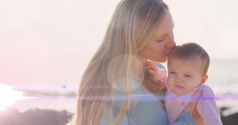 A mother gently kisses her baby while holding them close, showcasing a bond filled with love and affection. Taken outdoors during sunset, the light creates a warm, glowing effect, adding a dreamy atmosphere. Suitable for campaigns related to Mother's Day, parenting, family lifestyle, and emotional connection themes.