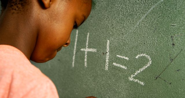 A young African American child is focused on solving a simple math problem written on a green chalkboard, with copy space. Capturing the essence of early education, the image reflects a moment of learning and concentration.