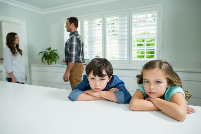 Two children, a boy and a girl, appear sad and distressed while leaning on a white surface. In the background, their parents are having an argument. This image can be used to depict family conflicts, emotional distress in children, and the impact of parental arguments on kids. Suitable for articles, blogs, and educational materials on family dynamics, mental health, and child psychology.