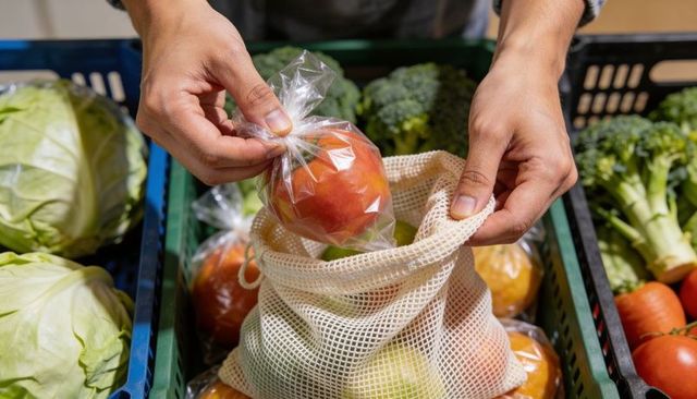 Shopper placing wrapped apple into reusable mesh bag at farmers market with fresh produce