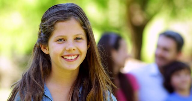 Young teenage girl smiling at camera while other family members are slightly blurred in the background in a park setting. Great for family-oriented advertisements, websites focusing on family activities, outdoor leisure, and youthful joy. Can be used for social media, brochures, and banners promoting family bonding or outdoor events.