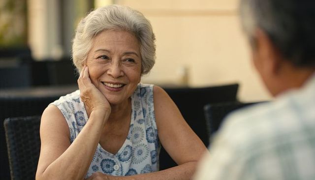 Smiling senior asian woman chatting on sunny patio leaning chin on hand