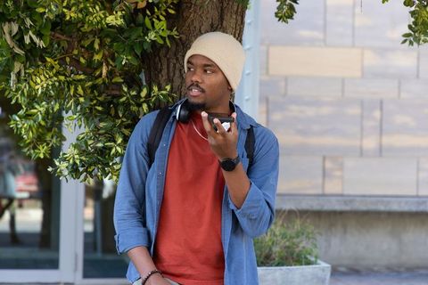 African American commuter leaning on tree speaking into smartphone wearing headphones
