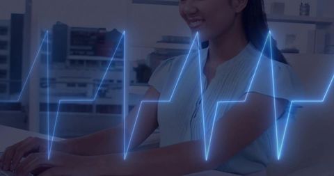 Smiling asian woman typing at desk with neon ecg waveform overlay, digital heartbeat