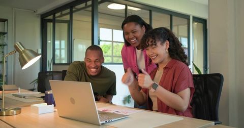Diverse team celebrating project win while collaborating on laptop in modern office