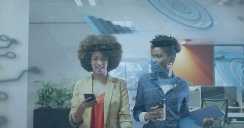 Two black professional women walking in modern office checking smartphone and holding coffee