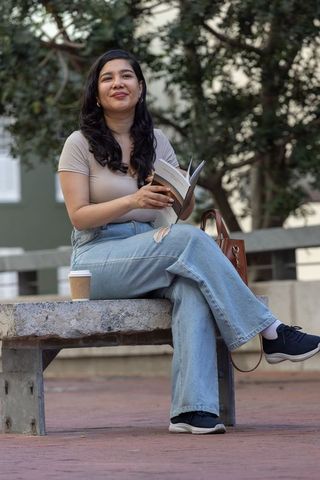 Young woman reading paperback on urban bench with coffee cup and leather tote in denim