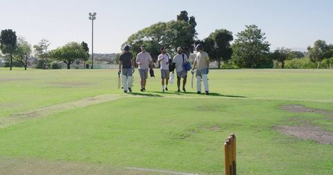 Cricket players walking across green pitch carrying bats and kit while chatting and laughing