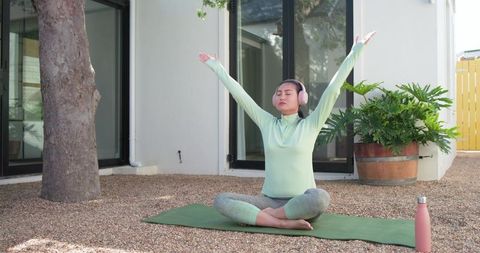 Asian woman practicing backyard meditation with headphones on green yoga mat for wellness