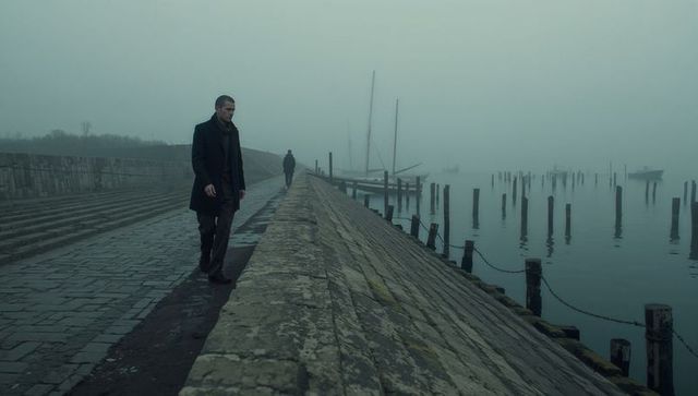 Walking man in dark overcoat along foggy cobbled quay with mooring posts and masts