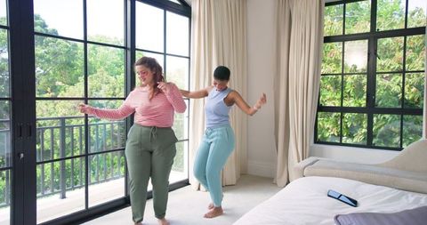 Friends dancing barefoot in sunlit modern bedroom by balcony, joyful morning routine