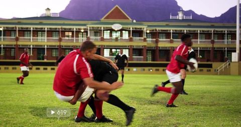 Rugby players tackling during match on grass field with historic school building backdrop