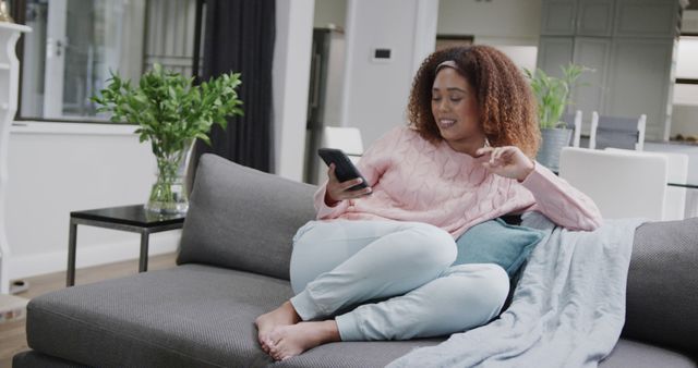 Young woman in cozy clothing sitting on a comfortable gray couch at home, looking at her smartphone with a happy expression. Ideal for depicting modern lifestyle, technology use, leisure time, home comfort, and social media engagement. Can be used in advertisements, lifestyle blogs, tech product promotions, and social media posts.