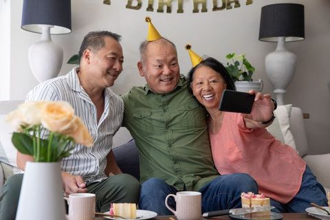 Asian family celebrating birthday together taking selfie on cozy sofa with cake and hats