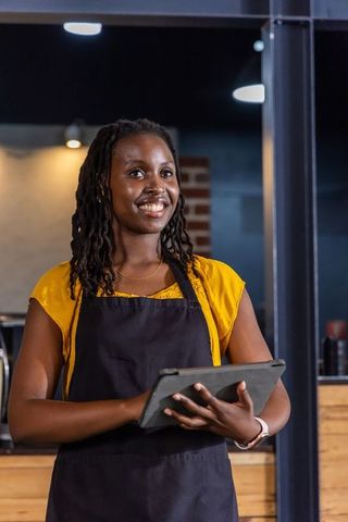 African american barista smiling while holding tablet behind modern coffeehouse counter