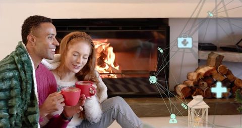 Couple enjoying cozy evening by fireplace sipping hot drinks under blanket with firewood